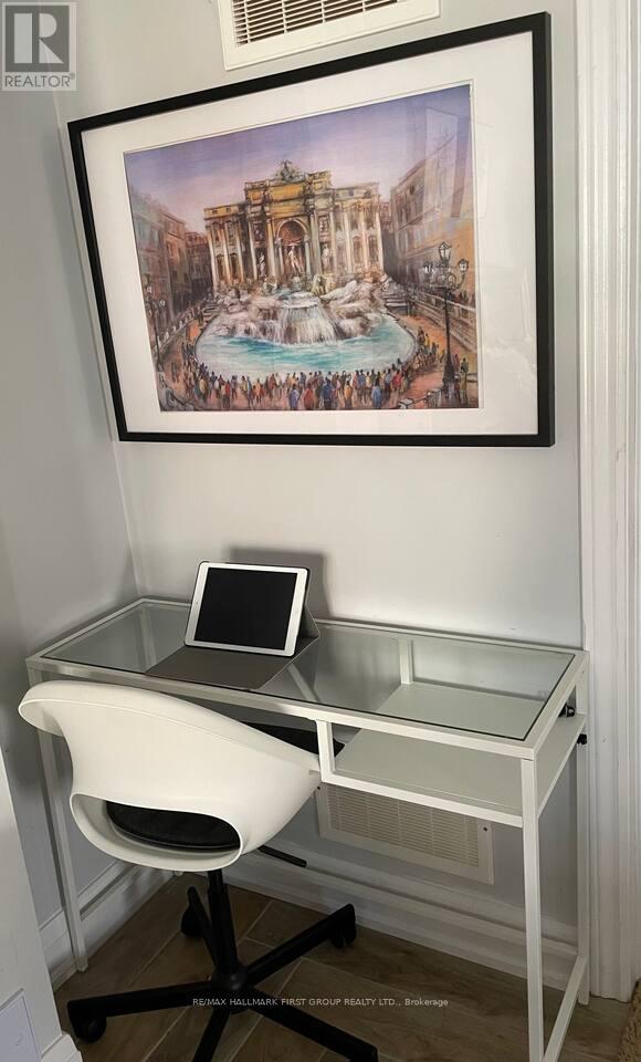 A framed picture of a fountain is hanging on a wall above a desk with a laptop and chair.