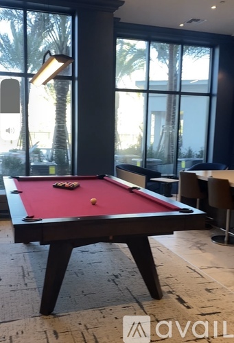A pool table with a red cloth and white balls is in the foreground of a room with large windows.