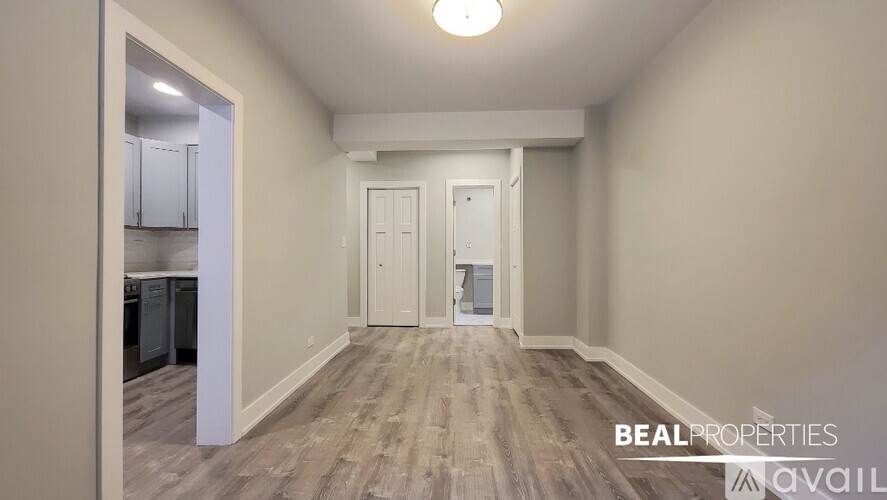 A spacious room with wooden flooring and white walls, with a kitchen visible through an open door.