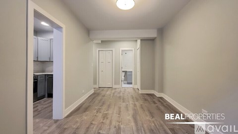 A spacious room with wooden flooring and white walls, with a kitchen visible through an open door.