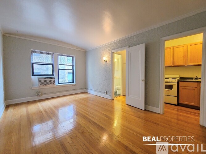 A room with wooden floors and a view of a kitchen through an open door.