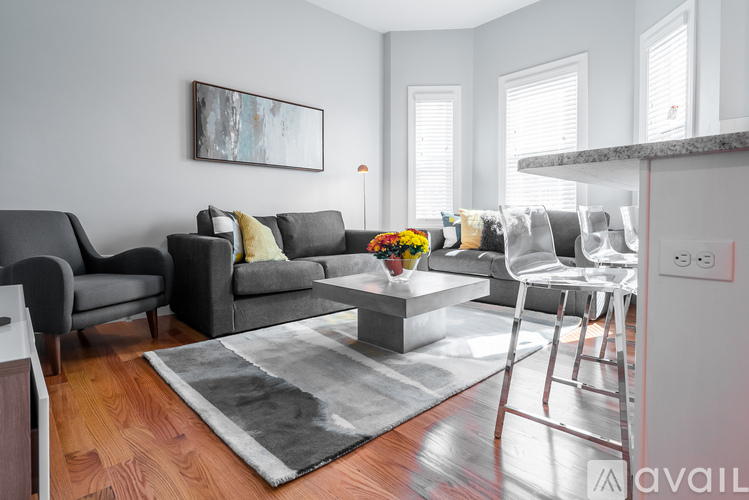 A living room with a grey sofa, a grey armchair, a grey chair, a grey coffee table, a grey rug, a painting on the wall, a window with blinds, a kitchen counter with a sink, a white fr