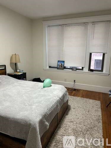 A bedroom with a bed, a nightstand with a lamp, a window with blinds, and a picture frame on the window sill.