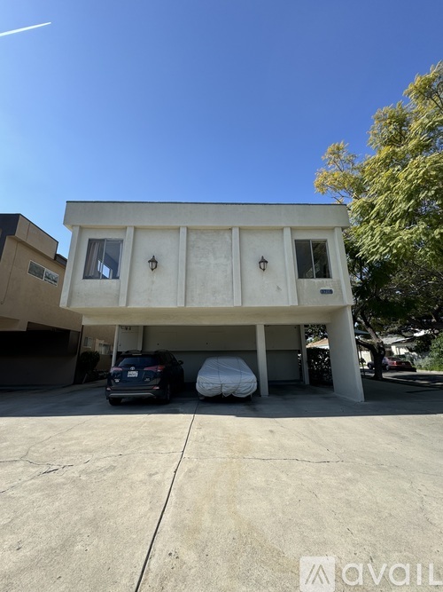 A car is parked in front of a white building with a tree to the right.