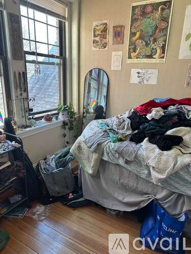 A messy bedroom with a bed covered in blankets and clothes, a window with a view of a street, and a wall with various posters and artwork.