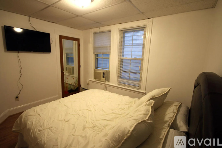 A bedroom with a bed, a window with blinds, and a flat-screen TV mounted on the wall.