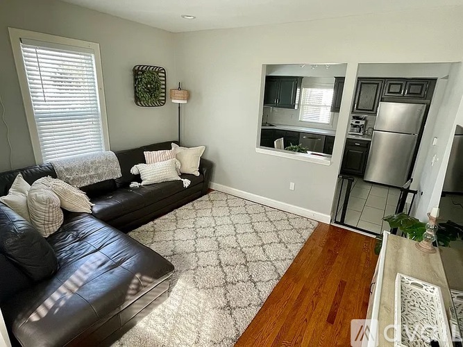 A black leather couch with white pillows sits in a living room with a kitchen in the background.