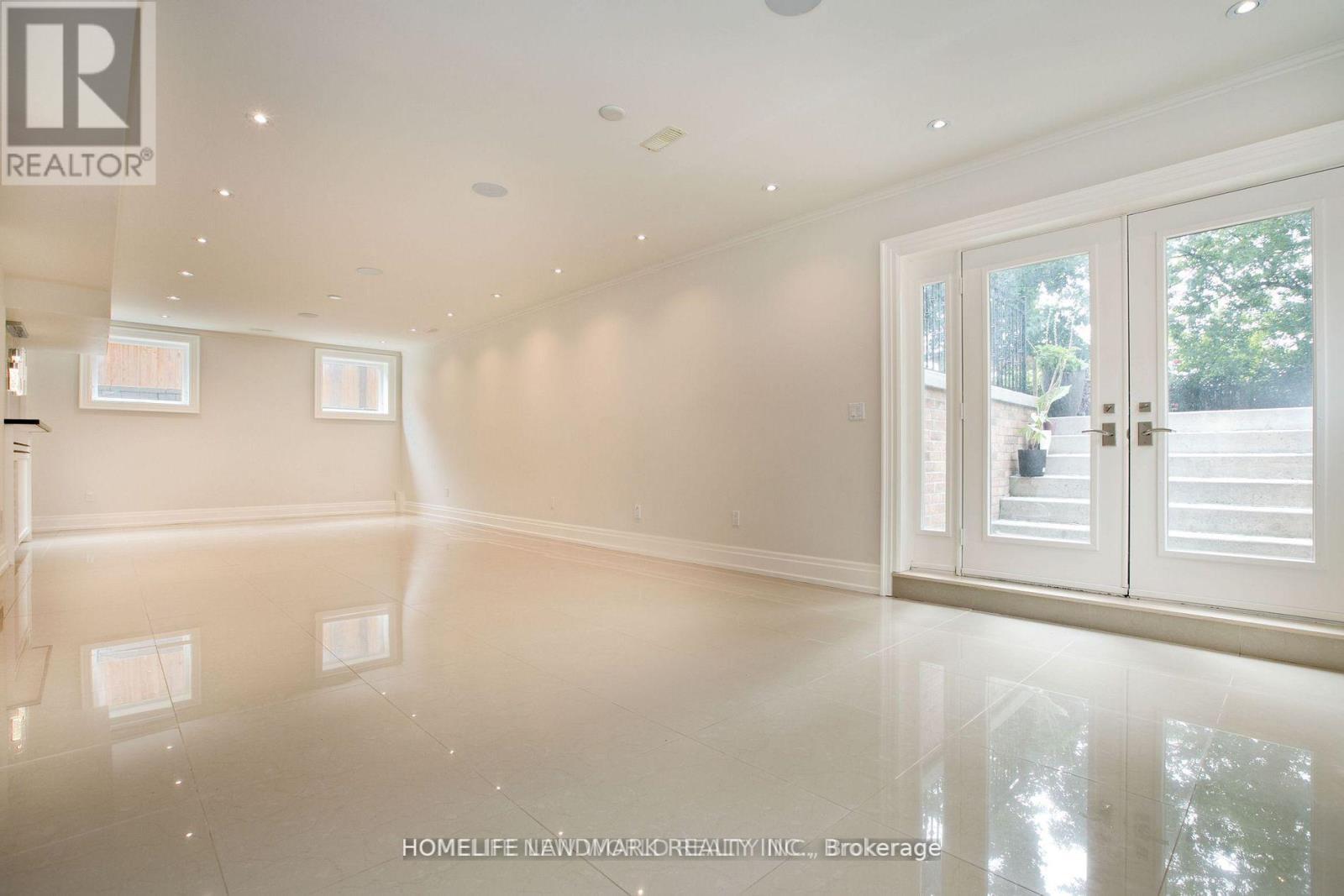 A spacious, empty room with white walls and flooring, and a large window with a view of trees outside.