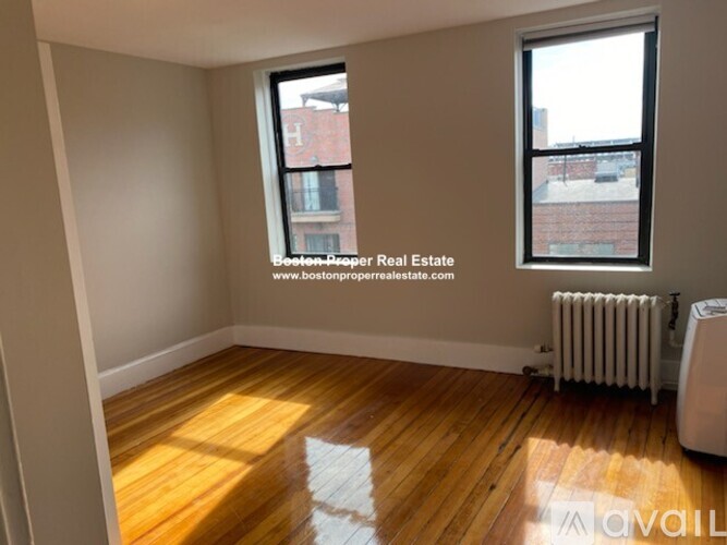 A room with wooden floors and two windows, one of which has a view of a red building.
