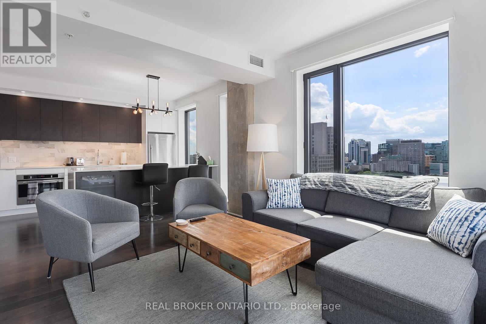 A modern living room with a grey sofa, a grey armchair, a wooden coffee table, and a city view through the window.