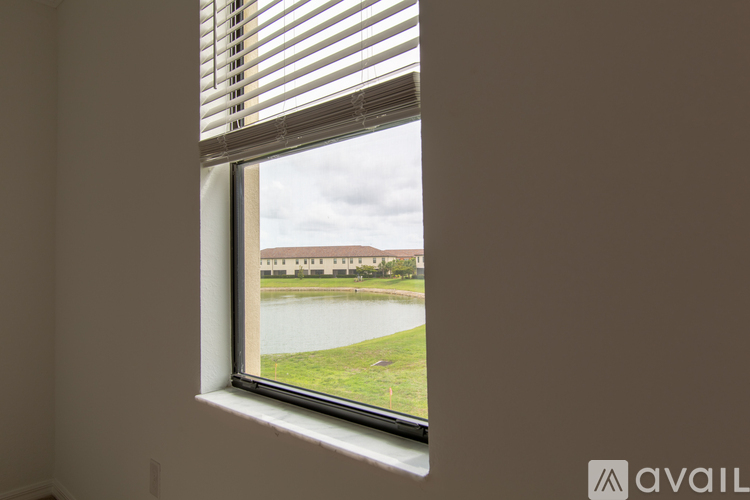 A window with blinds overlooking a green field and buildings.