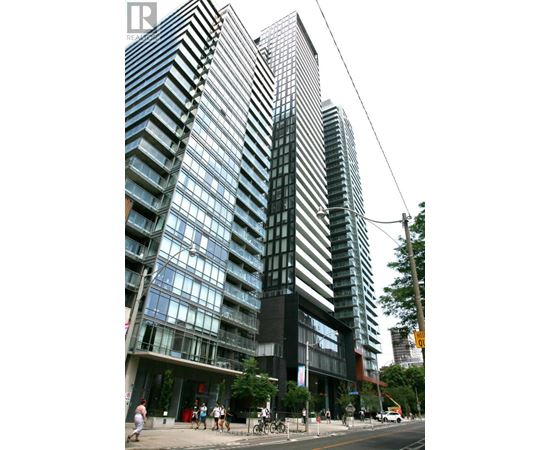 A tall building with a glass facade stands next to a street with people walking on the sidewalk.