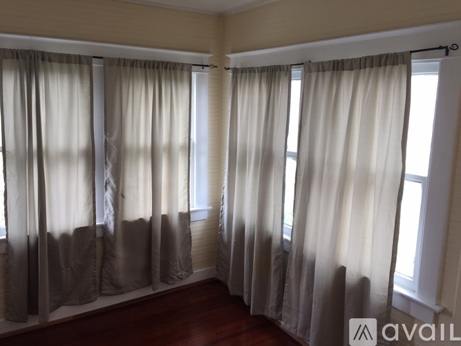 A room with beige walls and a wooden floor with curtains hanging on the windows.