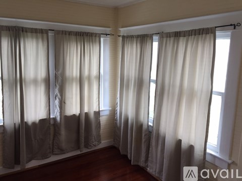 A room with beige walls and a wooden floor with curtains hanging on the windows.