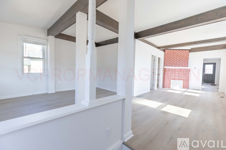A spacious room with white walls and wooden flooring, featuring a large window and a fireplace with a brick surround.