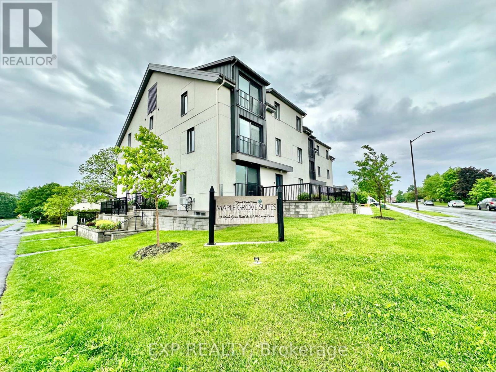 A modern two-story building with a balcony on the second floor is surrounded by a grassy area and a street in front.