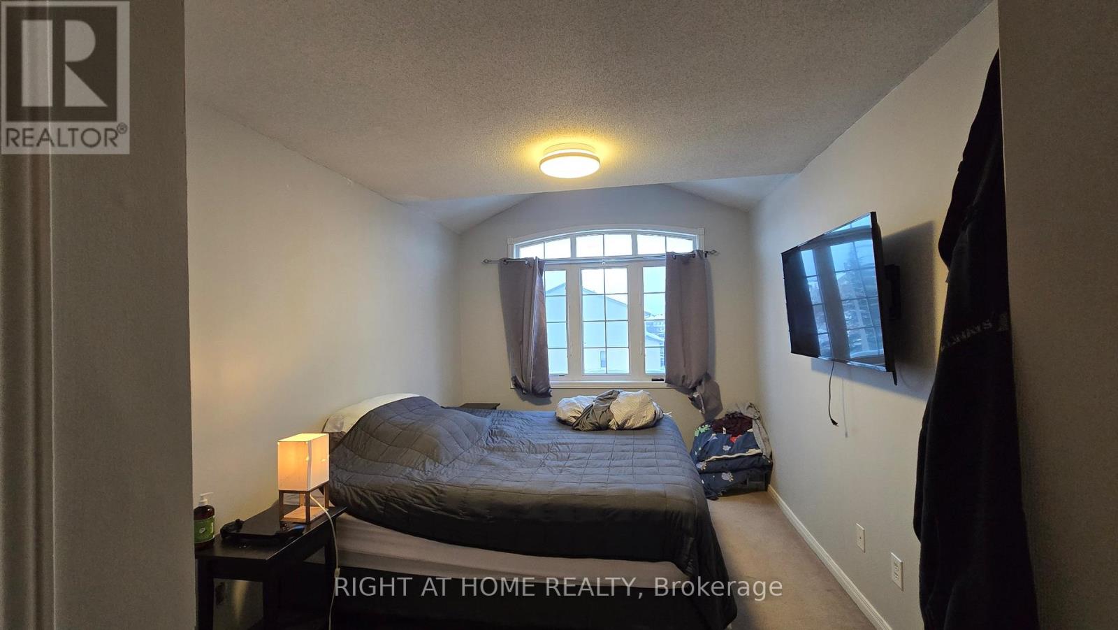 A bedroom with a bed, a nightstand, a window with curtains, and a flat-screen TV mounted on the wall.