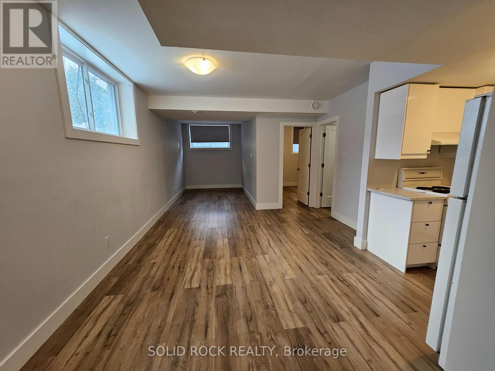 A spacious room with wooden flooring and white walls, furnished with a desk and a refrigerator.