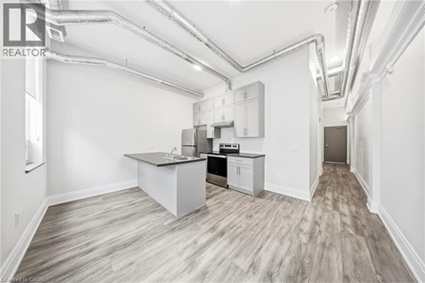 A spacious kitchen with a sink, stove, and cabinets in a white room with wood flooring and exposed pipes.