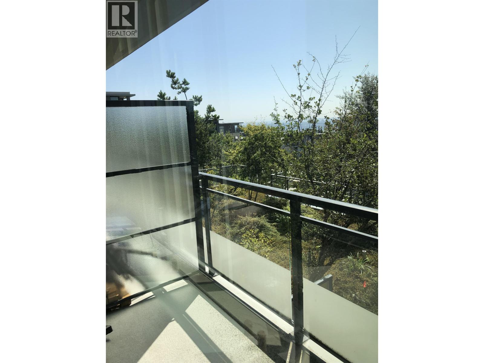 A glass balcony with a metal railing and a view of trees and buildings.