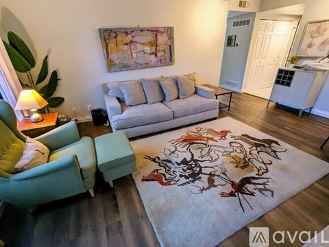 A living room with a grey couch, a green armchair, a yellow chair, a rug with a design on it, a painting on the wall, a lamp on a side table, and a white door in the background.