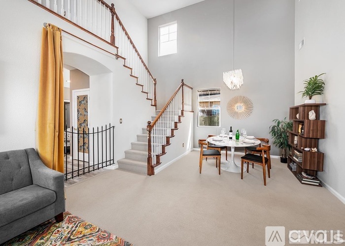 A living room with a grey couch, a dining table with chairs, a staircase with a railing, and a potted plant.