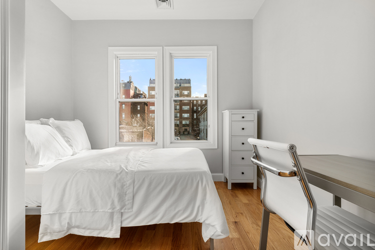 A bedroom with a white bed, a white chair, and a window with a view of a cityscape.