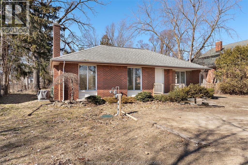 A house with a brown brick exterior and a grey roof is surrounded by a dirt lot.