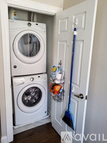 A white washing machine is in a small space next to a door with a mop and a basket of cleaning supplies.