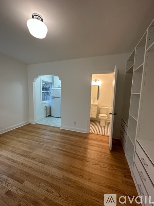 A room with wooden floors and white walls, with a kitchen visible through an open door.