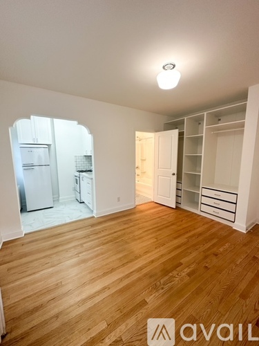 A room with wooden floors and white walls, with a kitchen visible through an arched opening.
