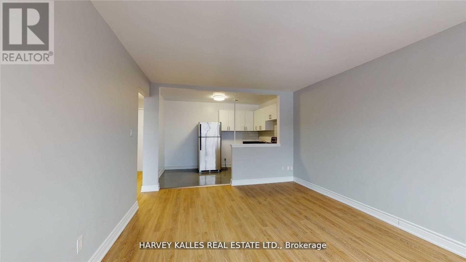 A spacious room with wooden flooring and white walls, with a kitchen area visible in the background.