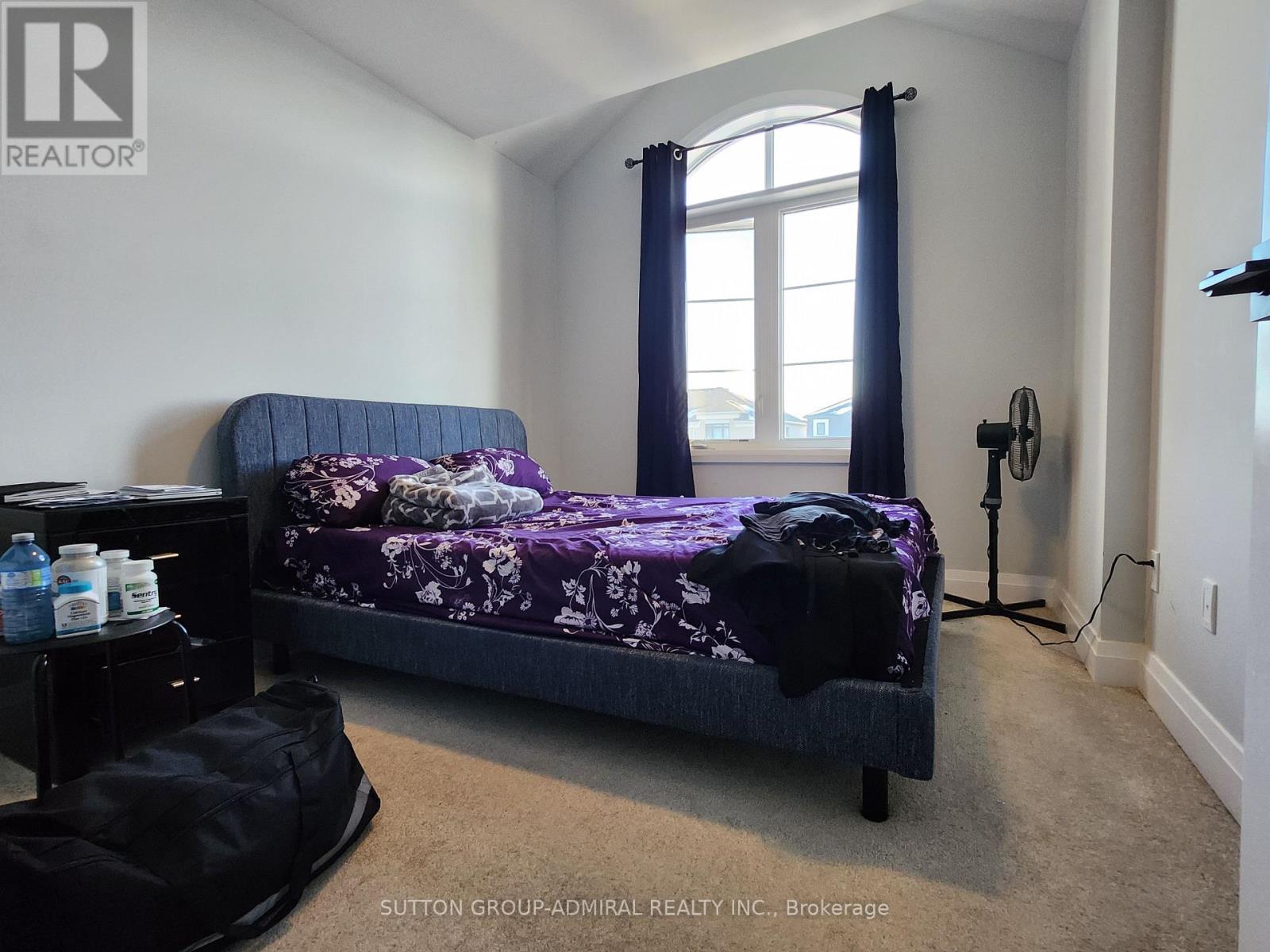 A bedroom with a bed, a window with curtains, a nightstand with bottles, and a black bag on the floor.