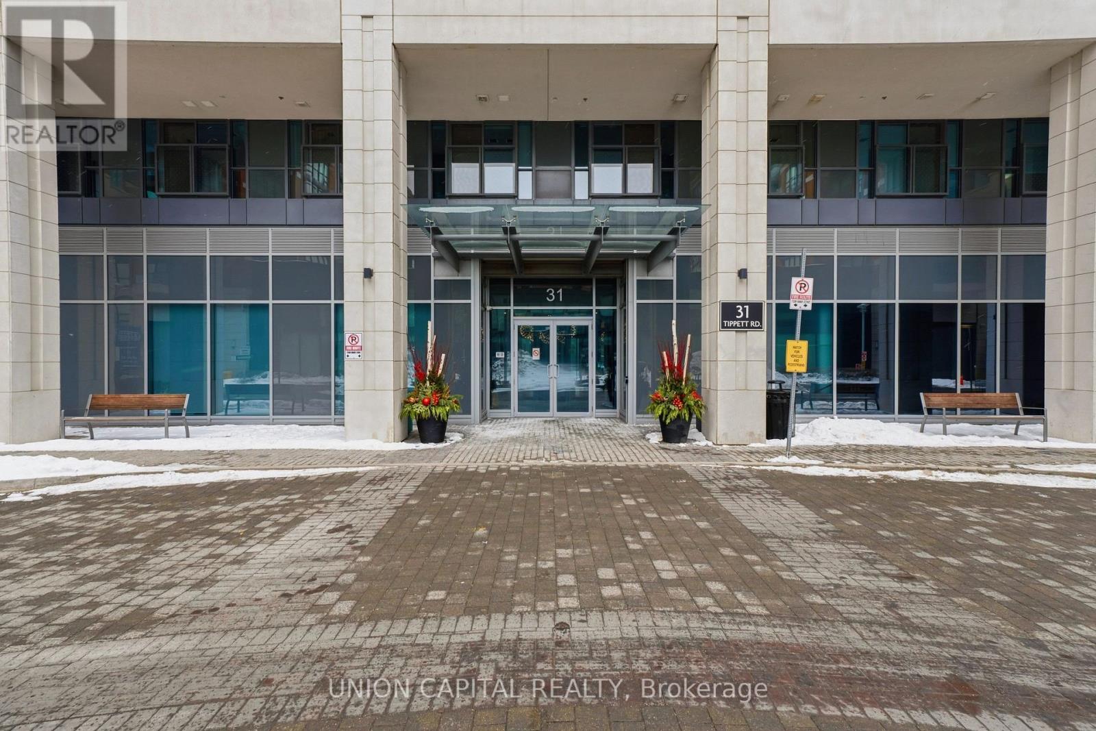 The image shows the front of a building with a glass entrance and a brick walkway leading to it.