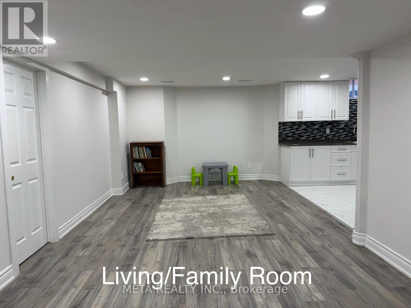 A living room with a rug, a bookshelf, and a kitchen area in the background.