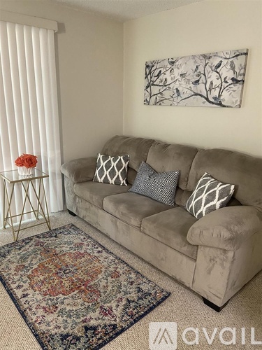 A living room with a beige couch, a small table with a red flower, a rug, and a painting on the wall.
