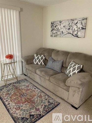 A living room with a beige couch, a small table with a red flower, a rug, and a painting on the wall.