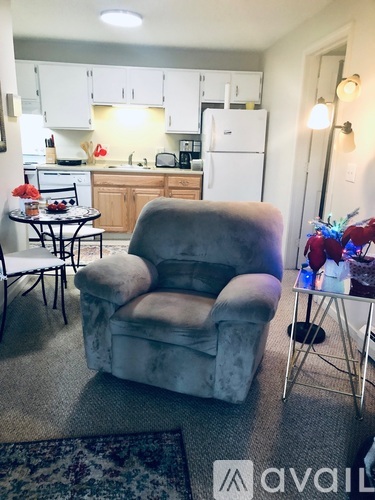 A grey recliner chair is in the middle of a carpeted room with a table and chairs in the kitchen area.
