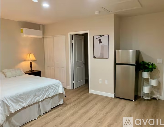 A bedroom with a bed, a lamp, a refrigerator, and a plant.