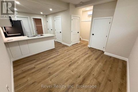 A room with wooden flooring and white walls, with a kitchen area on the left and a door on the right.