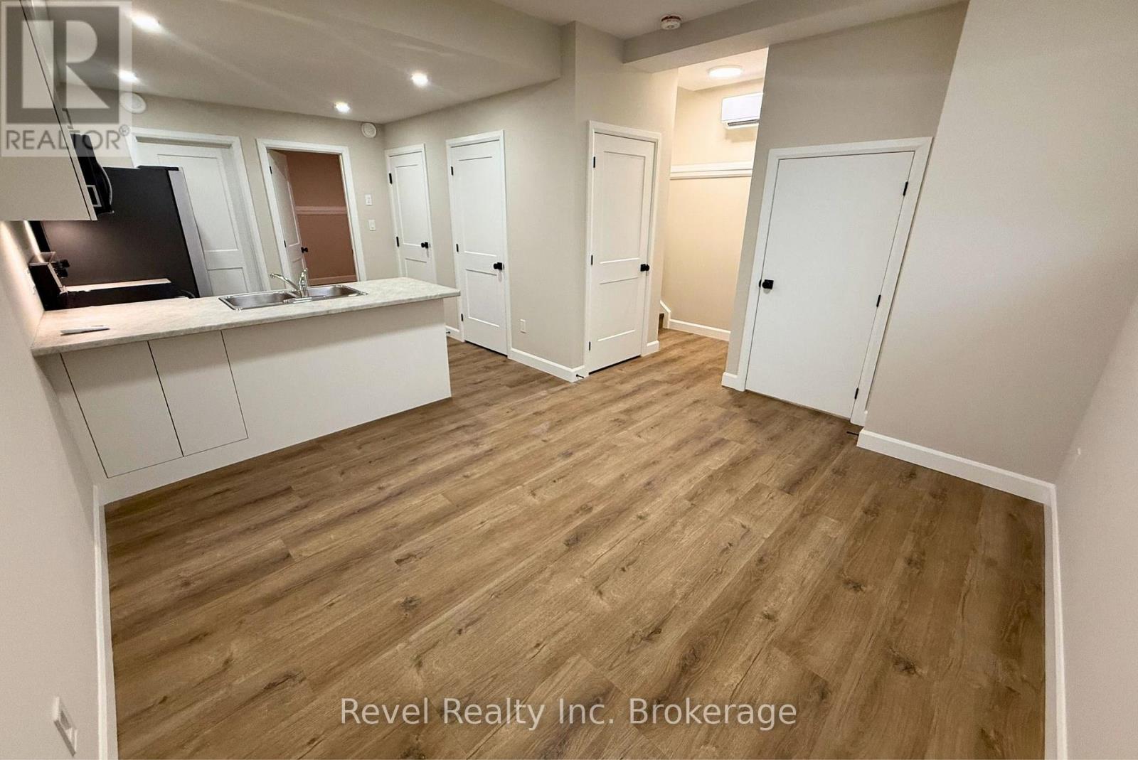 A room with wooden flooring and white walls, with a kitchen area on the left and a door on the right.