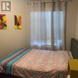 A bed with a patterned comforter sits in a bedroom with a window covered by sheer curtains.