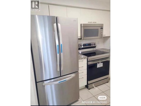 A stainless steel refrigerator stands in a kitchen with a microwave and oven in the background.