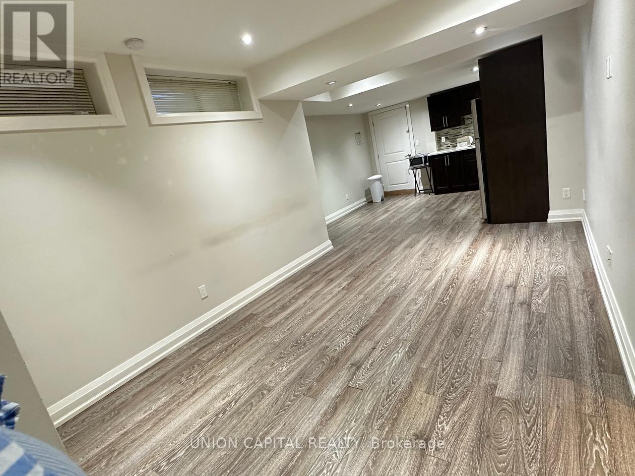 A spacious room with wood flooring and a kitchen area in the background.