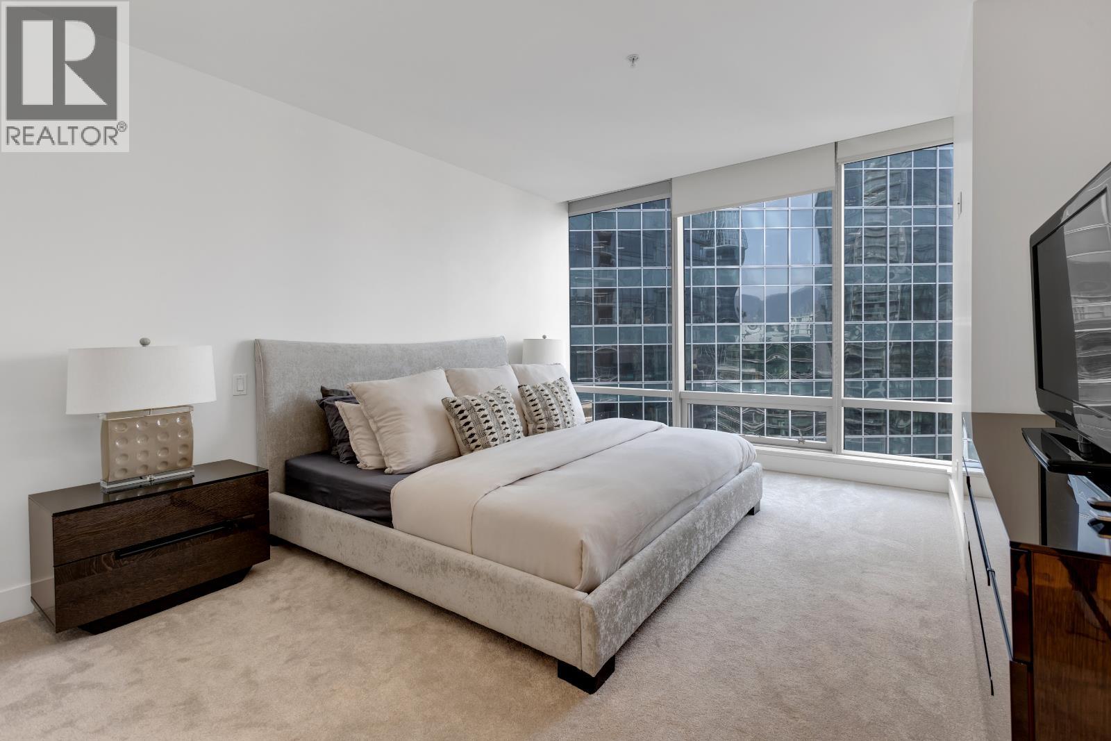 A bedroom with a large bed, a nightstand with a lamp, and a window with a view of the city.