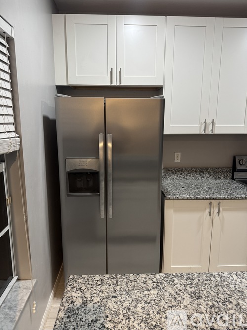 A stainless steel refrigerator with a water and ice dispenser in the middle of the door stands in a kitchen with white cabinets.