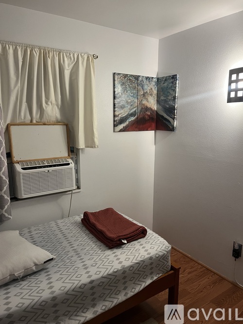 A bedroom with a bed, a fan, a painting, and a suitcase.