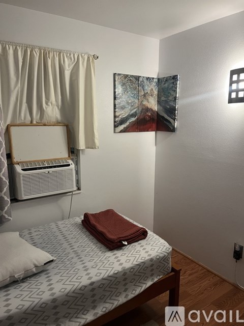 A bedroom with a bed, a fan, a painting, and a suitcase.