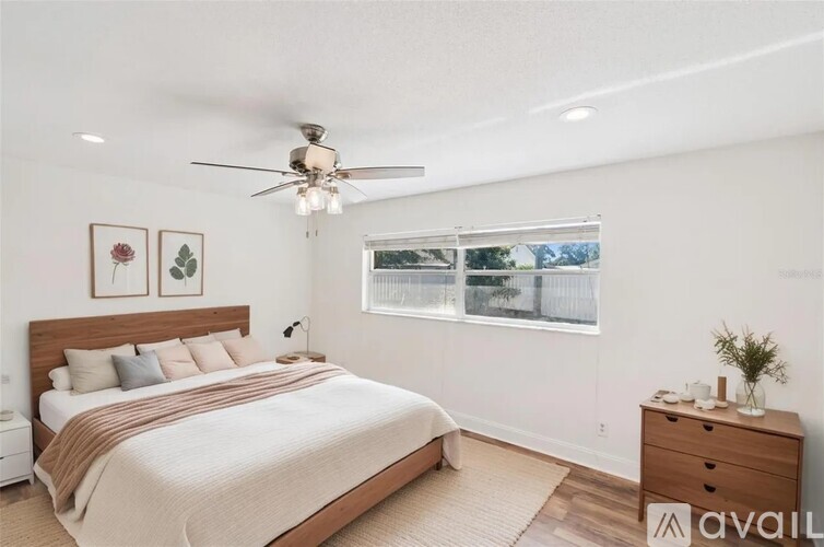 A bedroom with a large bed, a ceiling fan, and a window with a view of a fence and trees.