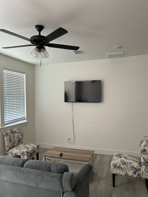 A living room with a grey couch, a chair with a floral pattern, a television on the wall, and a ceiling fan with lights.